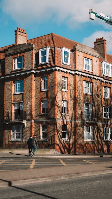 Exterior view of a four-story brick building with several white-framed windows, located on a street with pavement and marked parking spaces. Shadows of leafless trees are cast onto the building's faca