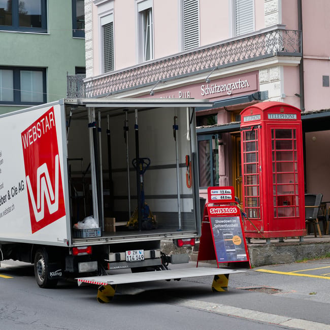A moving truck with a partially open side panel is parked on a street in front of a residential building, revealing an interior containing packing materials and equipment such as a hand truck and straps. The truck is positioned on the road, with stabilising wheel chocks visible at the rear. Nearby, a red traditional British telephone box stands on the pavement next to an advertising board for Swisslos. The building behind features pastel-colored facades, window shutters, and a balcony with decorative wrought iron railings. The scene captures elements related to house removals and furniture transport, illustrating the logistics involved in a home relocation process, with a focus on loading goods onto a moving vehicle in an urban environment. The lighting suggests daytime conditions, suitable for recording a typical scene of pack-and-move activities, supported by Kennington Man and Van as a professional removals service provider.