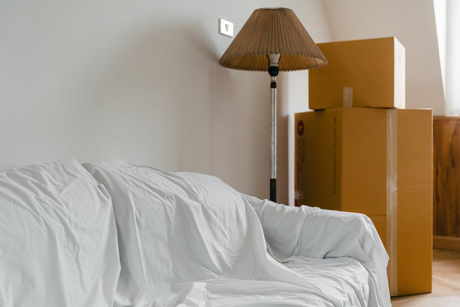 In a domestic interior scene, three cardboard moving boxes of varying sizes are stacked horizontally on a wooden furniture piece, with the largest at the bottom, the medium-sized boxes in the middle, and a smaller box on top. To the left, a white sofa draped with a cloth is partially visible, and behind the boxes, a tall floor-standing lamp with a brown pleated lampshade provides ambient lighting. The wall in the background is light-colored, and a window with partially drawn curtains allows natural light into the room. The image captures the process of home relocation, with packed boxes prepared for moving and furniture wrapped for transport, reflecting a typical house removal setting supported by Kennington Man and Van's professional services.