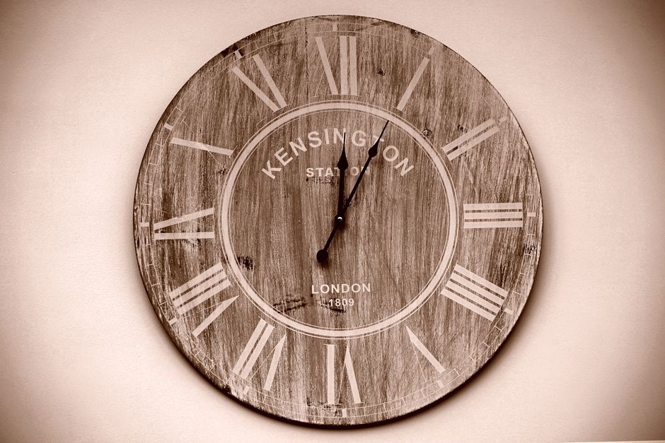 A round, vintage-style wall clock with a wooden face and white Roman numerals. The clock displays the time as approximately 12:03 and features the text 'KENSINGTON' and 'LONDON 1809' in the center. It
