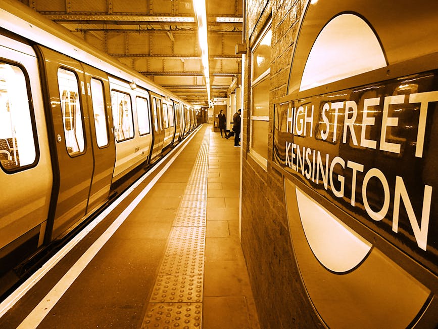 A round, vintage-style wall clock with a wooden face and white Roman numerals. The clock displays the time as approximately 12:03 and features the text 'KENSINGTON' and 'LONDON 1809' in the center. It