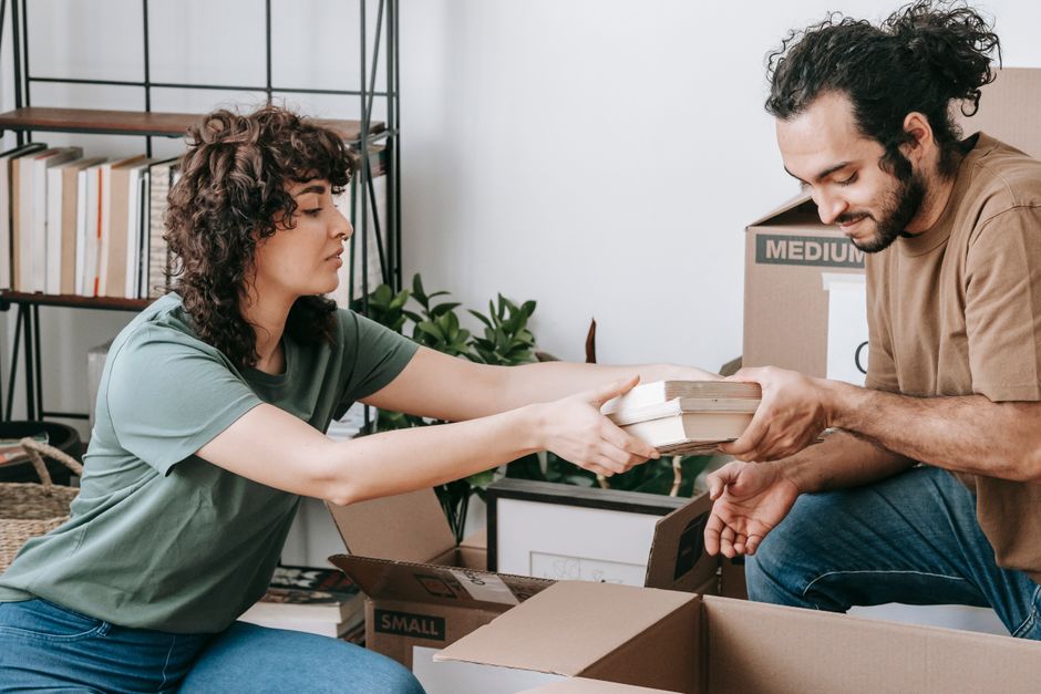A woman with curly brown hair and wearing a green t-shirt is inside a home, reaching out to receive a stack of books wrapped in white paper from a man with dark, curly hair and a beard, dressed in a brown t-shirt. They are in a room with shelving units filled with books and plants, surrounded by cardboard boxes, some labeled for packing and moving. The man is kneeling on the floor near an open box, assisting with packing or unpacking during a home relocation process. The scene reflects packing and moving logistics relevant to house removals, with visible packaging materials like plastic wrap and cardboard, and furniture such as a bookshelf and a potted plant, all under natural indoor lighting.