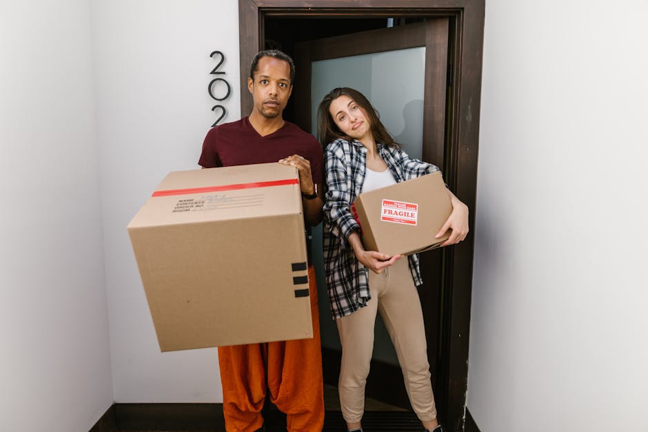 A man and a woman stand in a narrow indoor hallway near a doorway, each holding a cardboard box for home relocation purposes. The man, dressed in a maroon shirt and orange trousers, is holding a large box with red and black tape, while the woman, wearing a plaid shirt over a white t-shirt and beige trousers, carries a smaller box marked with a fragile sticker. The background features a white wall with the number '200' visible and a dark wooden door frame surrounding a closed door. The flooring appears to be dark-colored vinyl or laminate, and natural lighting illuminates the scene. This image depicts the careful handling of packed belongings during furniture transport and the loading process involved in professional removals, highlighting the importance of packing durability and proper handling to ensure a smooth removal experience, as emphasized by services like Kennington Man and Van.
