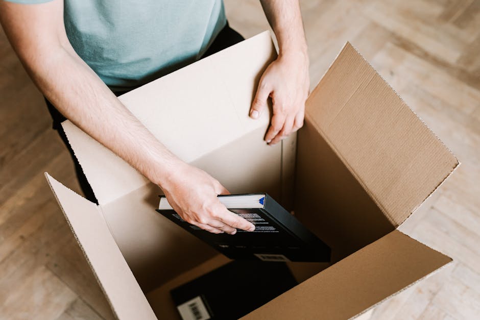 A woman with curly brown hair and wearing a green t-shirt is inside a home, reaching out to receive a stack of books wrapped in white paper from a man with dark, curly hair and a beard, dressed in a brown t-shirt. They are in a room with shelving units filled with books and plants, surrounded by cardboard boxes, some labeled for packing and moving. The man is kneeling on the floor near an open box, assisting with packing or unpacking during a home relocation process. The scene reflects packing and moving logistics relevant to house removals, with visible packaging materials like plastic wrap and cardboard, and furniture such as a bookshelf and a potted plant, all under natural indoor lighting.