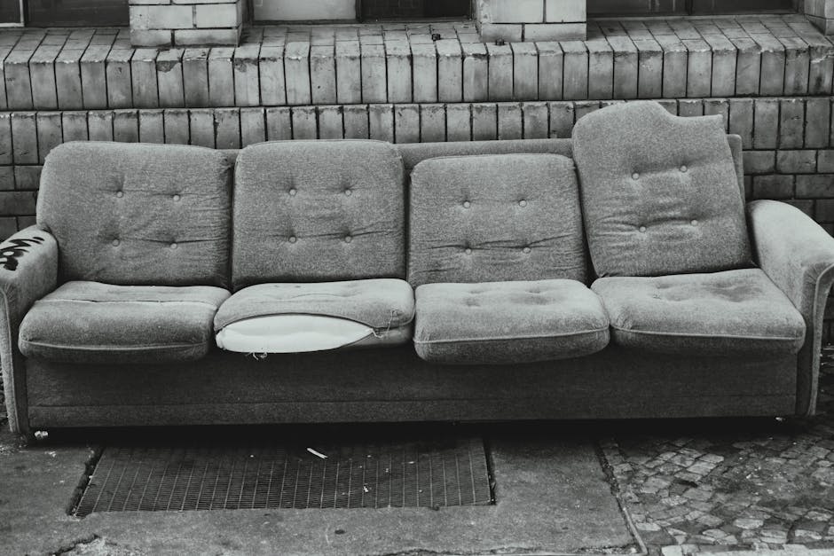 A black and white photograph of an old, worn-out three-seater fabric sofa positioned outdoors against a brick wall. The sofa's cushions show visible signs of wear, with a torn section on the middle seat revealing the padding inside, and the fabric appears wrinkled and slightly stained. The sofa has padded armrests on both ends, with the left armrest featuring a small label or sticker partially visible. It is situated on a paved surface with a metal drainage grate directly beneath it, suggesting a sidewalk or street setting. Behind the sofa, there is a neatly stacked brick wall, indicating an urban environment. The setting appears to be part of a home relocation or furniture transport process, with the sofa possibly awaiting removal by a professional moving service such as Kennington Man and Van involved in house removals or furniture disposal, supported by the context of house removal services and local regulations for furniture disposal in Lambeth.