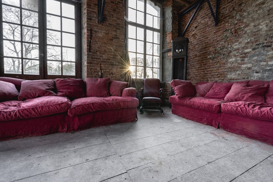 A spacious room with exposed brick walls and large industrial-style windows allowing natural sunlight to fill the space. In the foreground, there are two large, plush sofas upholstered in deep pink fabric, each with matching cushions, positioned facing each other. A dark wooden armchair with a high back and rolled arms is situated between the sofas, closer to the windows. The floor is made of wide, light-colored wooden planks, with some visible dust and wear. In the corner to the right, a black freestanding stove or heater is present, with a chimney pipe extending upward. Visible lifting straps and packing materials such as cardboard boxes and plastic wraps are not present in this image, but the scene appears to be prepared for a home relocation process involving furniture transport. The lighting highlights the textures of the brick walls and fabric upholstery, implying a professional removal company's involvement, like Kennington Man and Van, in the furniture moving or disposal process as part of house clearance or moving services.