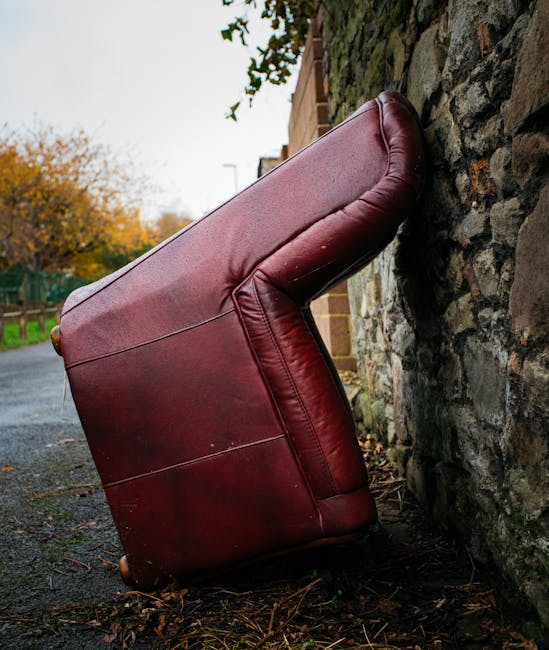 A black and white photograph of an old, worn-out three-seater fabric sofa positioned outdoors against a brick wall. The sofa's cushions show visible signs of wear, with a torn section on the middle seat revealing the padding inside, and the fabric appears wrinkled and slightly stained. The sofa has padded armrests on both ends, with the left armrest featuring a small label or sticker partially visible. It is situated on a paved surface with a metal drainage grate directly beneath it, suggesting a sidewalk or street setting. Behind the sofa, there is a neatly stacked brick wall, indicating an urban environment. The setting appears to be part of a home relocation or furniture transport process, with the sofa possibly awaiting removal by a professional moving service such as Kennington Man and Van involved in house removals or furniture disposal, supported by the context of house removal services and local regulations for furniture disposal in Lambeth.