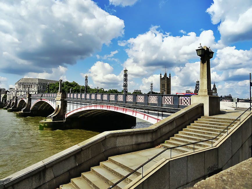 A photograph captured during daytime showing Westminster Bridge spanning the River Thames in central London. The bridge features ornate lamp posts, decorative railings, and stone arches supporting its structure. The river below has a greenish-brown hue, with gentle water flow visible. In the background, the iconic Elizabeth Tower, known as Big Ben, stands tall with its Gothic architecture, alongside the Houses of Parliament, partly obscured by trees and a partly cloudy sky. Visible on the right side of the image are stone steps with a metal handrail leading down from the bridge, adjacent to a paved walkway. The scene is well-lit with natural sunlight, highlighting the architectural details and the general setting associated with city centre London. This image relates to house removals and relocation services in the area, possibly indicating the need for legal permits or logistical planning for property moves near Westminster. The atmosphere suggests a busy urban environment with historic landmarks, suitable for a service provider like Kennington Man and Van involved in furniture transport, packing, and home relocation within the borough.