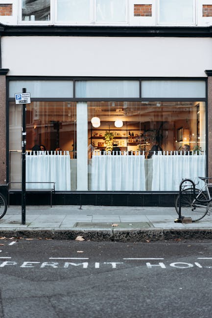 The image shows a ground-floor commercial space with large glass windows and a sliding glass door, revealing the interior of a café or restaurant illuminated by warm lighting. The front exterior features a narrow sidewalk with two bicycles secured to metal bike racks on either side of the entrance. The sidewalk borders a street with street markings, including part of a designated parking area. The building's exterior is primarily dark-colored with a white signboard above the windows. Inside, visible elements include tables, chairs, decorative plants, and shelves with various items, all fitted with curtains that partially obscure the view. The scene is captured during daylight, with natural light illuminating the sidewalk and street. Though the image does not depict house removals or transport activities directly, it exemplifies residential or commercial relocation logistics, fitting within the service scope of Kennington Man and Van for house removals or furniture transport in the Kennington area, as referenced in the Lambeth Council permit guide for large moves.