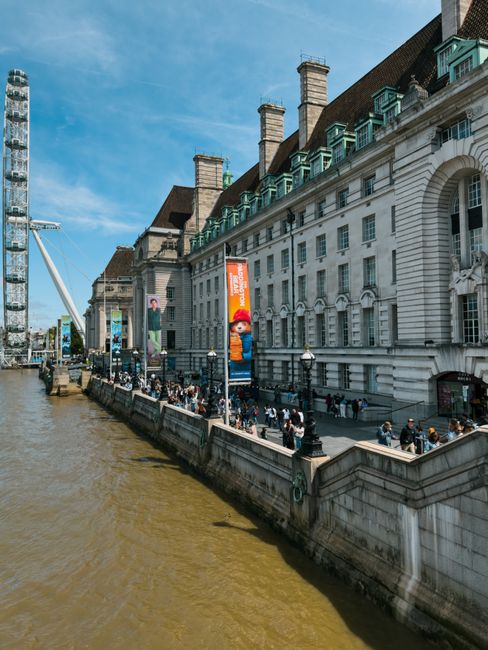 A photograph captured during daytime showing Westminster Bridge spanning the River Thames in central London. The bridge features ornate lamp posts, decorative railings, and stone arches supporting its structure. The river below has a greenish-brown hue, with gentle water flow visible. In the background, the iconic Elizabeth Tower, known as Big Ben, stands tall with its Gothic architecture, alongside the Houses of Parliament, partly obscured by trees and a partly cloudy sky. Visible on the right side of the image are stone steps with a metal handrail leading down from the bridge, adjacent to a paved walkway. The scene is well-lit with natural sunlight, highlighting the architectural details and the general setting associated with city centre London. This image relates to house removals and relocation services in the area, possibly indicating the need for legal permits or logistical planning for property moves near Westminster. The atmosphere suggests a busy urban environment with historic landmarks, suitable for a service provider like Kennington Man and Van involved in furniture transport, packing, and home relocation within the borough.