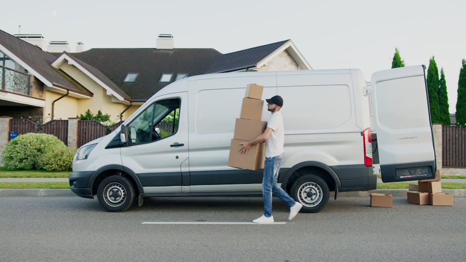 A person wearing a white shirt, blue jeans, and white sneakers walks along a suburban street next to a white panel van used by Kennington Man and Van for home relocation services. The individual is carrying three medium-sized cardboard boxes stacked vertically, with additional boxes placed on the pavement beside the van. The van's rear sliding door is open, indicating an active loading or unloading process, with some boxes close to the vehicle on the roadside. Behind the scene, there are modern residential houses with pitched roofs, large windows, and balconies, surrounded by well-maintained greenery including bushes and trees. The scene is set during daylight hours with a clear sky, illustrating a typical furniture transport or packing and moving scenario as part of house removals or relocation services.