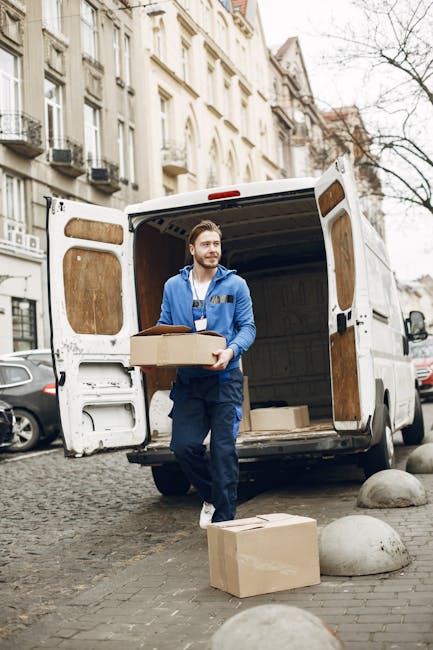 A male removal worker with a beard, wearing a blue jacket and navy overalls, stands outside a white cargo van with open rear doors on a cobblestone street in an urban residential area. He is carrying a cardboard box, and there are additional cardboard boxes on the ground nearby, some on the pavement and others being loaded into the van. The van's interior is visible, showing a spacious cargo area suited for home relocation and furniture transport. Surrounding the scene are parked cars, multi-storey residential buildings with balconies and large windows, and a leafless tree, indicating an outdoor setting during colder months. The lighting is natural, suggesting it is daytime, and the scene depicts the process of packing and moving items typical of professional removals carried out by companies like Kennington Man and Van.
