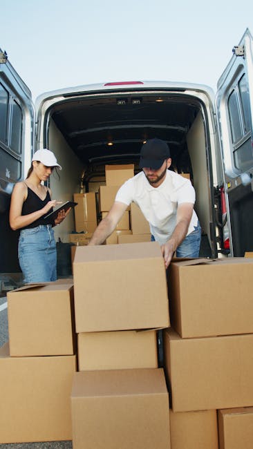 A person wearing a white shirt, blue jeans, and white sneakers walks along a suburban street next to a white panel van used by Kennington Man and Van for home relocation services. The individual is carrying three medium-sized cardboard boxes stacked vertically, with additional boxes placed on the pavement beside the van. The van's rear sliding door is open, indicating an active loading or unloading process, with some boxes close to the vehicle on the roadside. Behind the scene, there are modern residential houses with pitched roofs, large windows, and balconies, surrounded by well-maintained greenery including bushes and trees. The scene is set during daylight hours with a clear sky, illustrating a typical furniture transport or packing and moving scenario as part of house removals or relocation services.