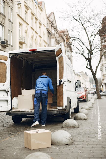 A man wearing a blue jacket and dark trousers is seen loading or unloading a large, open white moving van parked on a city street. The van's interior is lined with wooden panels and contains several c