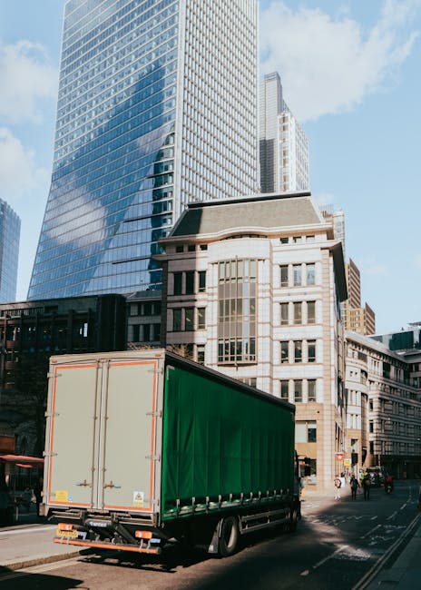A man wearing a blue jacket and dark trousers is seen loading or unloading a large, open white moving van parked on a city street. The van's interior is lined with wooden panels and contains several c