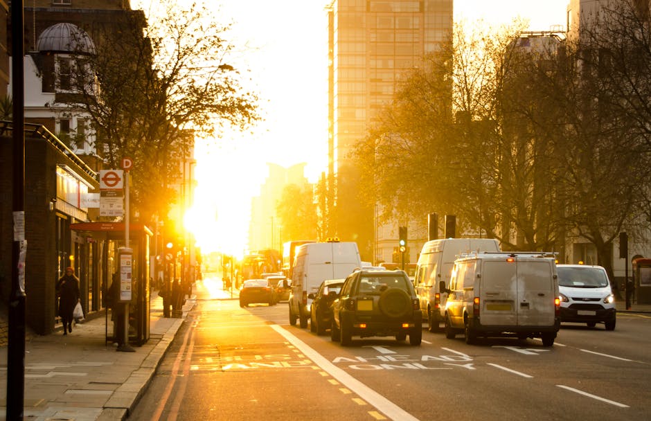 A city street at sunset with a busy loading process for a house removal, featuring several white and dark-colored vans parked along the curb, some with open rear doors indicating active loading of goods. The scene includes a sidewalk with pedestrians, a traditional brick building with a bus stop shelter, and tall modern office buildings in the background. Trees lining the street have shed some leaves, suggesting an autumn setting. The warm golden light from the setting sun reflects off the vehicles and buildings, creating an atmospheric glow. This environment illustrates the logistics of packing and moving items during a home relocation, supported by professional removals services such as those provided by Kennington Man and Van, especially highlighting the vehicle-based transport process involved in furniture and box delivery within urban settings.