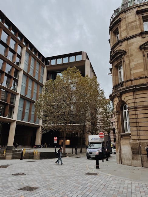 A city street scene showing the exterior of a modern office building with large glass windows and a concrete facade on the left, and a historic stone building with ornate architectural details and rounded bay windows on the right. In the middle ground, a large deciduous tree with sparse, light-colored leaves is visible, indicating autumn. The street is paved with light-colored bricks, and a few pedestrians are walking across the area. A white delivery van with branding parked near the stone building is part of the loading process, with a person seen standing beside it, possibly involved in furniture or box loading related to house or office removals. The scene is captured during daytime with overcast skies, and there are traffic bollards and a few traffic signs, including a 'no entry' sign, in the background. The image reflects typical urban logistics involved in professional moving and packing services, in line with [COMPANY_NAME]’s scope of house removals and furniture transport.