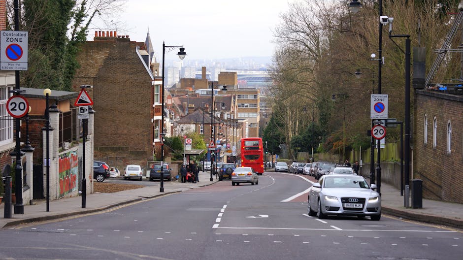 A residential street scene during daytime showing parked cars lining both sides of the narrow road, with various vehicles including vans and small trucks used for home relocation and furniture transpo